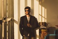 A fashionable man wearing sunglasses and a suit leans confidently against a sunlit wall.